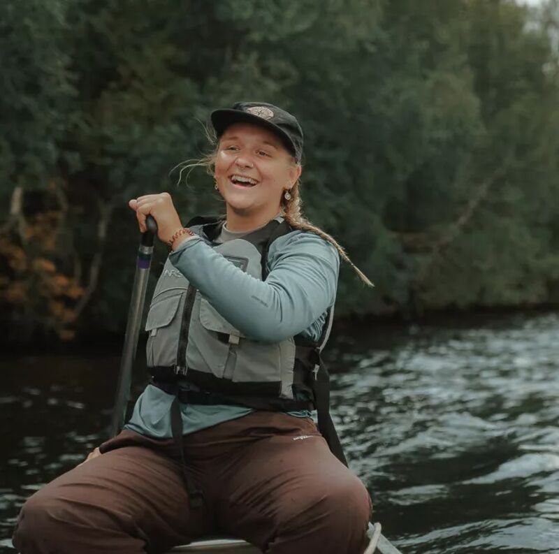 A woman wearing a cap, a light blue long-sleeved shirt, and a life vest is sitting in a canoe on a body of water. She is holding a paddle and smiling, appearing to be enjoying her time outdoors. The background features lush green trees, suggesting she is paddling in a natural setting, possibly a lake or river. The water is slightly choppy, indicating a gentle breeze or current.